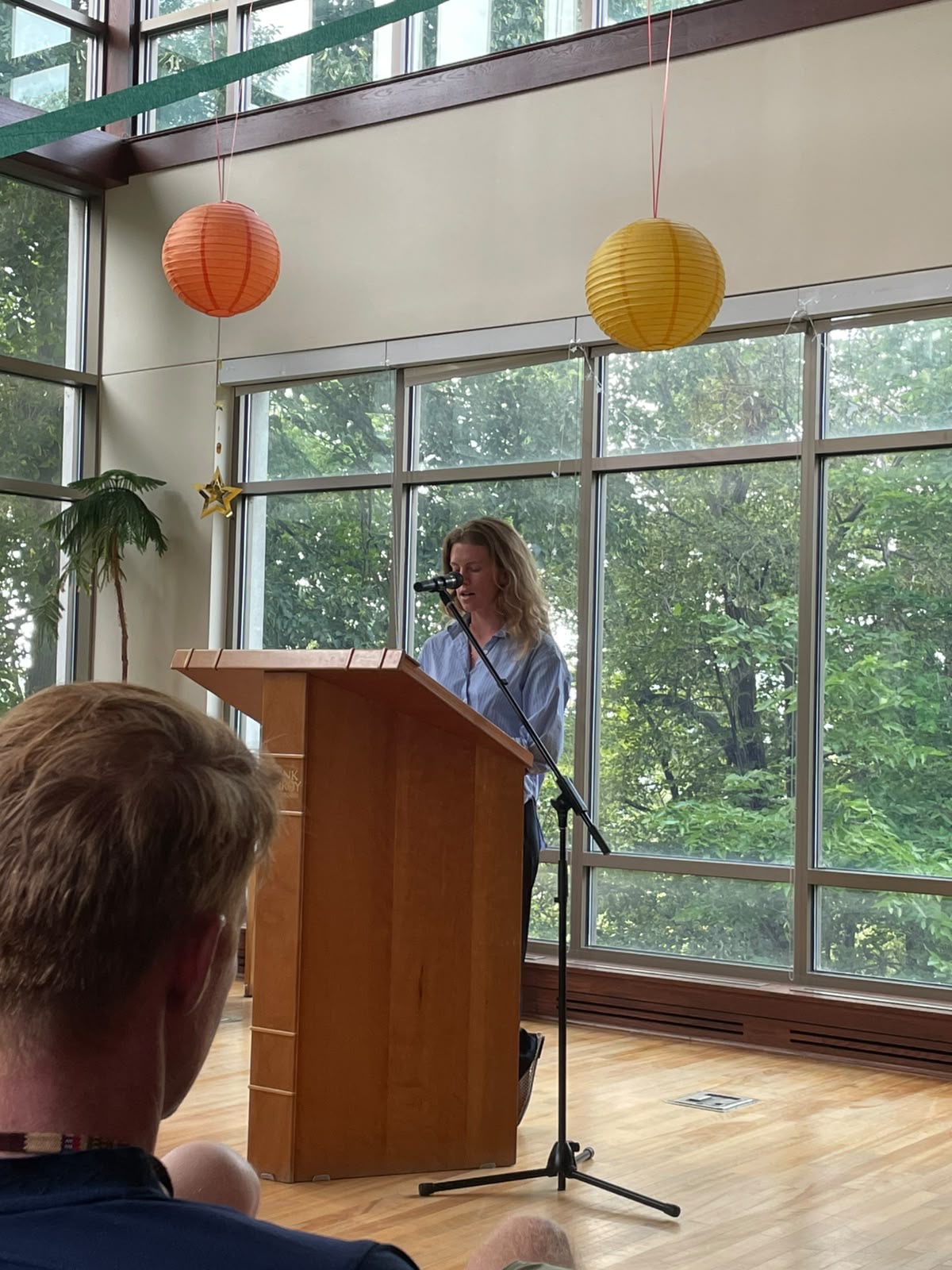 A writer reading at a podium beside tall windows